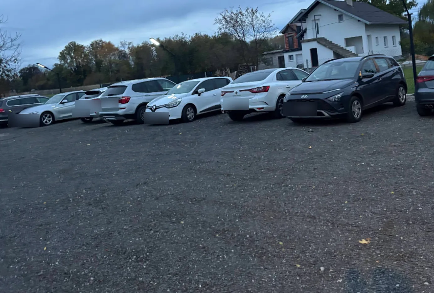 Stvarna fotografija parkinga danju: red parkiranih auta na šljunku uz rasvjetne stupove i obiteljsku kuću